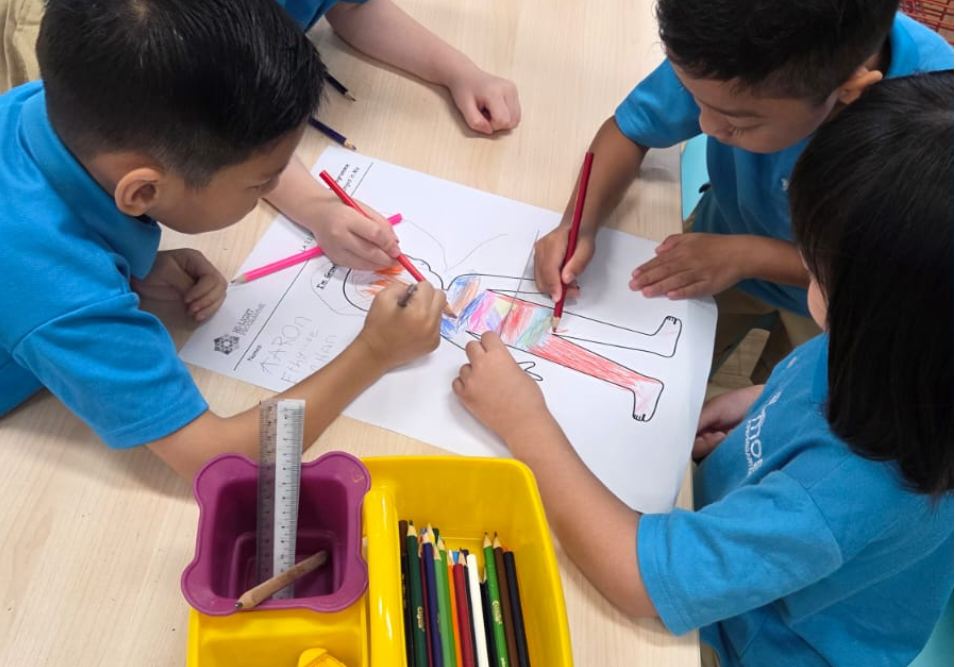 Four children in blue shirts color a large outline of a person on paper at a table with pencils.